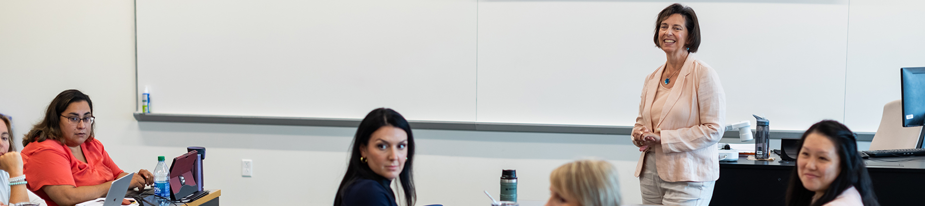 A female instructor stands at the front of a classroom and smiles at a group of adult students.